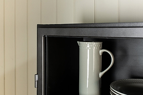 A white ceramic pitcher with a textured rim stands upright inside the nkuku Takua Iron Cabinet - Narrow, next to a stack of plates. Only the upper left section of the cabinet is shown against a cream-colored, paneled wall.