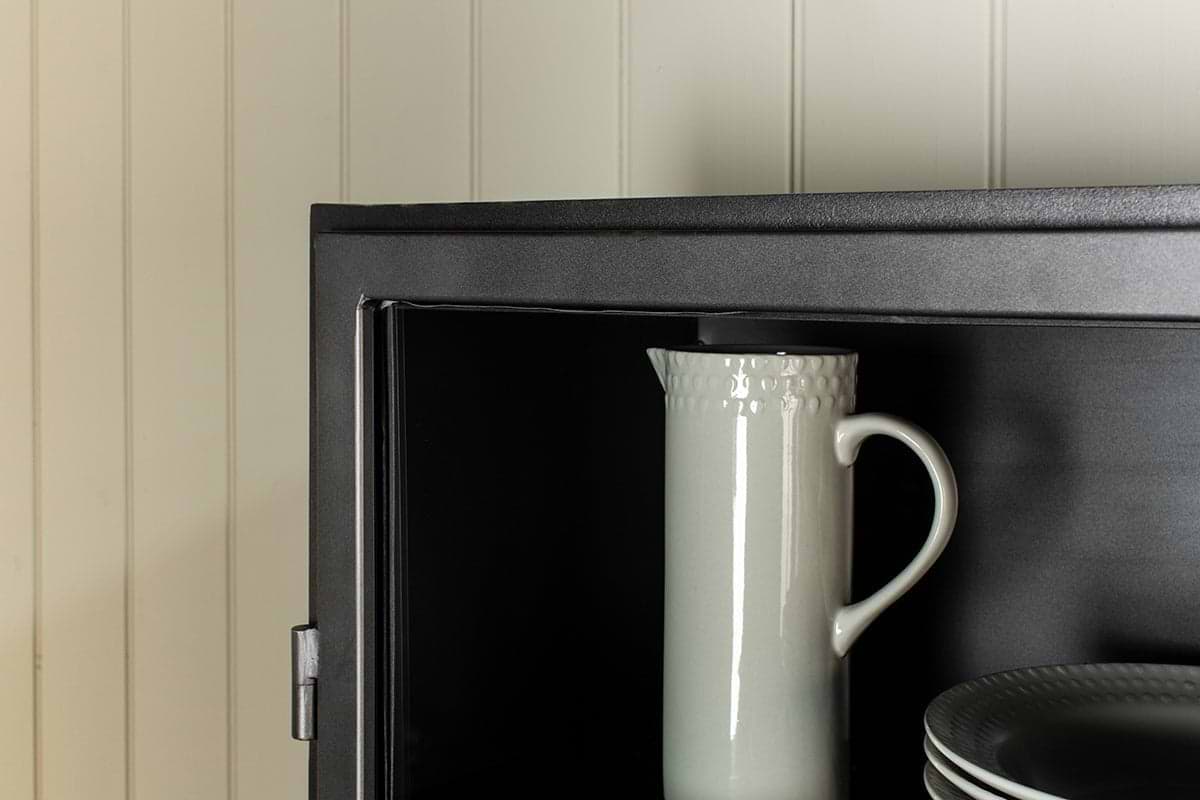 A white ceramic pitcher with a textured rim stands upright inside the nkuku Takua Iron Cabinet - Narrow, next to a stack of plates. Only the upper left section of the cabinet is shown against a cream-colored, paneled wall.