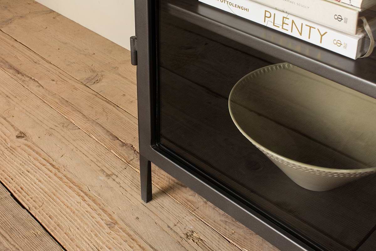 The Takua Iron Cabinet - Wide by nkuku showcases a beige bowl on a dark wooden shelf behind its glass door, with books arranged above—offering a minimalist blend of style and function.