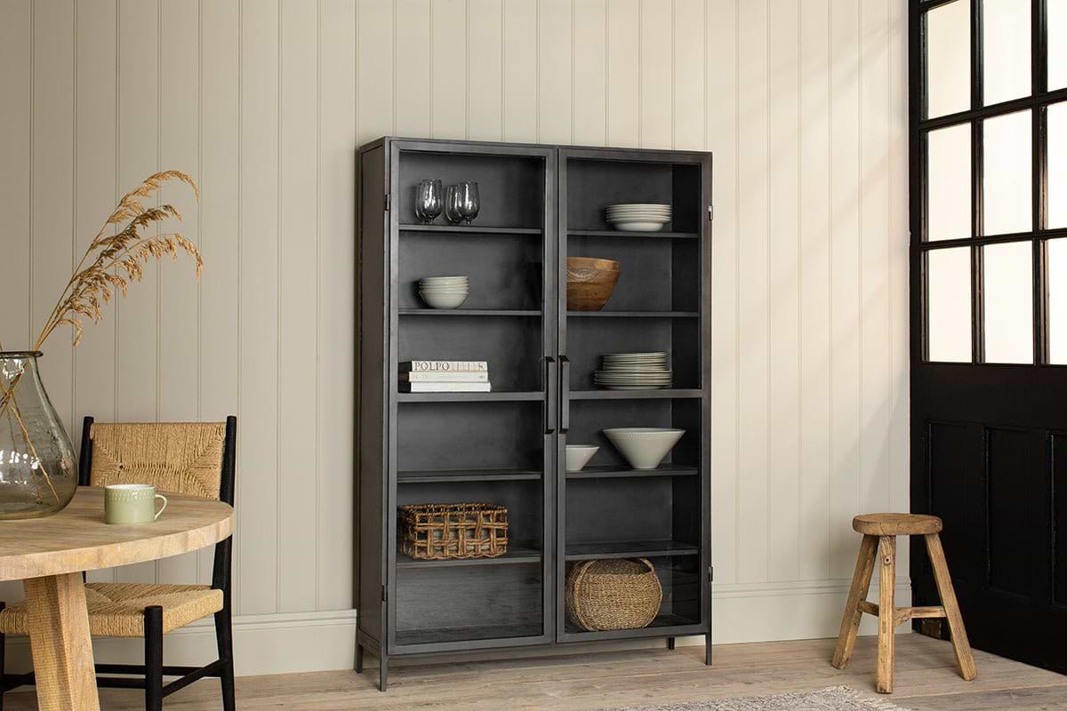 The Takua Iron Cabinet - Wide by nkuku, with a black glass front, stands against a cream paneled wall displaying dishes, bowls, glasses, books, and baskets. A round wooden table and woven chair sit nearby in the sunlit, cozy room.
