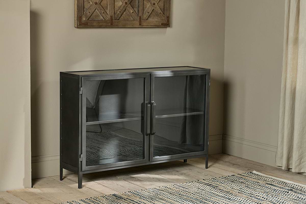 The Takua Iron Sideboard by nkuku, with glass doors and two shelves, stands against a beige wall on a light wooden floor. A woven rug is partially visible below, with a curtain to the right and rustic wooden décor mounted above.