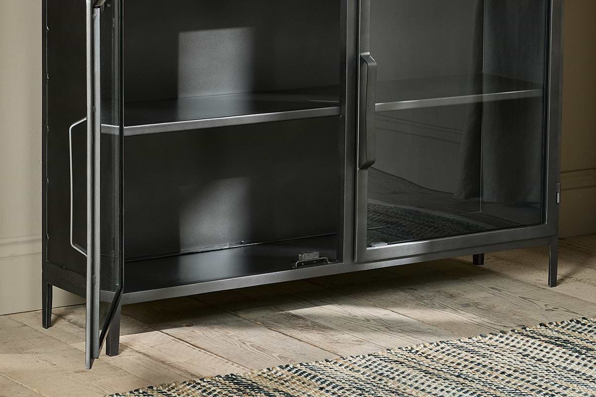 A close-up of the nkuku Takua Iron Sideboard, an industrial black metal and glass cabinet with two glass doors—one ajar—showing two shelves inside. The sideboard stands on four legs on a light wooden floor beside a cream and dark blue woven rug.