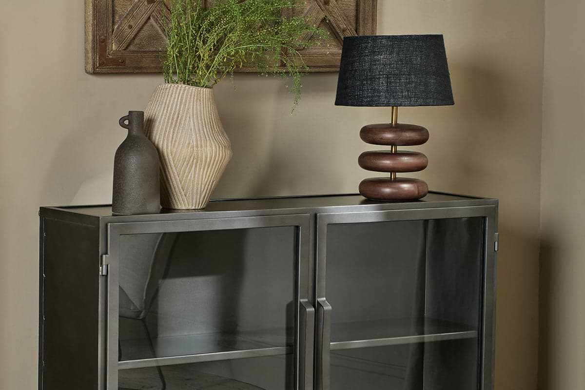 The Takua Iron Sideboard by nkuku stands against a beige wall, topped with a tall beige textured vase of green foliage, a smaller brown bottle-shaped vase, and a table lamp featuring a black shade and sculptural wooden base.