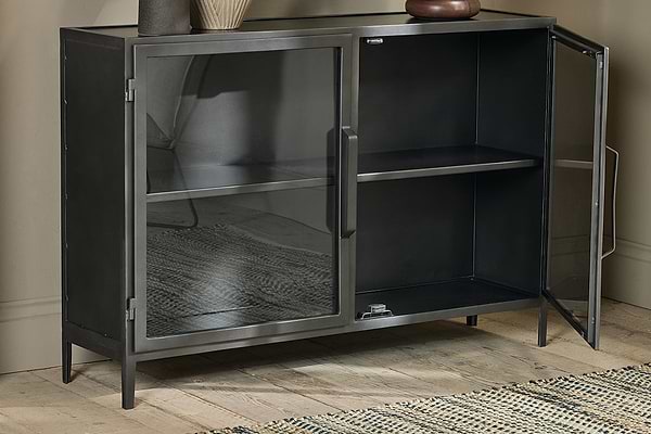The Takua Iron Sideboard by nkuku, featuring an industrial black metal design and two glass doors, stands on wooden flooring against a beige wall. It has a single interior shelf, with a vase and bowl on top and a woven rug partially visible on the left.