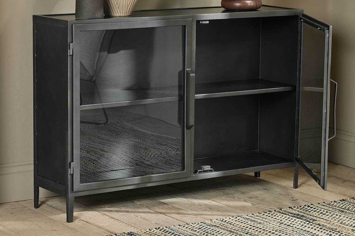 The Takua Iron Sideboard by nkuku, featuring an industrial black metal design and two glass doors, stands on wooden flooring against a beige wall. It has a single interior shelf, with a vase and bowl on top and a woven rug partially visible on the left.