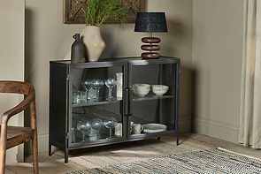 The nkuku Takua Iron Sideboard displays glassware and white dishes behind a glass front. On top are two vases—one ribbed with greenery, one dark and narrow—and a black-shaded modern lamp, alongside a wood chair and patterned rug.