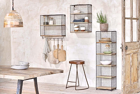 A rustic kitchen features nkuku’s Tamba Shelf with Hooks holding dishes, bowls, and plants. Wooden boards and towels hang from the hooks. A rust-finished shelving unit stands by the door, and a wooden table sits beneath a pendant lamp.
