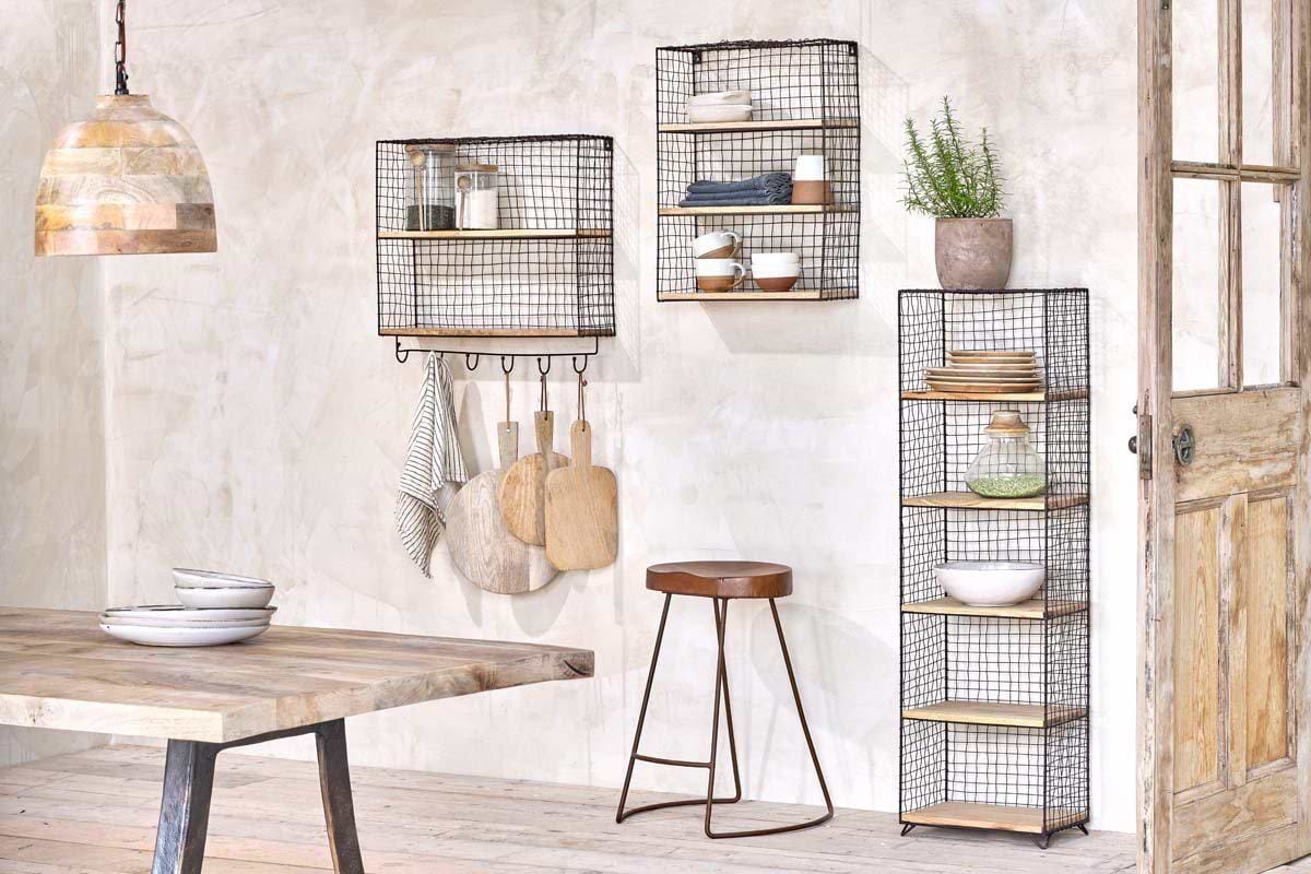 A rustic kitchen features nkuku’s Tamba Shelf with Hooks holding dishes, bowls, and plants. Wooden boards and towels hang from the hooks. A rust-finished shelving unit stands by the door, and a wooden table sits beneath a pendant lamp.