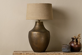 The nkuku Tanda Etched Statement Table Lamp in antique brass with a wide base and beige shade sits on a rustic wooden surface alongside two small pottery vases—one brown with dried flowers, one gray—against a neutral wall.