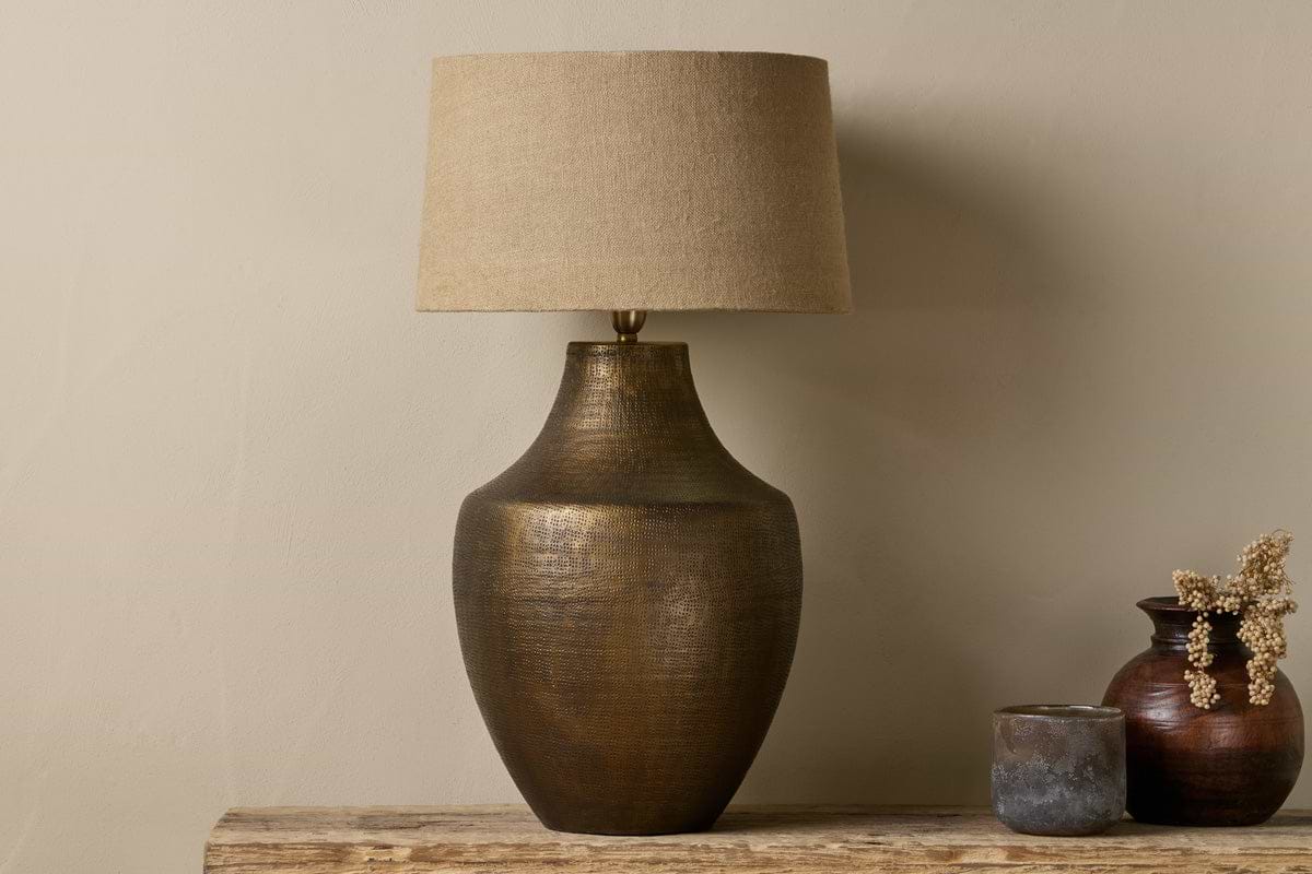 The nkuku Tanda Etched Statement Table Lamp in antique brass with a wide base and beige shade sits on a rustic wooden surface alongside two small pottery vases—one brown with dried flowers, one gray—against a neutral wall.