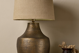 A close-up of the nkuku Tanda Etched Statement Table Lamp in Antique Brass (Extra Large) with a textured finish and beige fabric shade, set against a neutral wall. A small vase with dried flowers stands to the right.