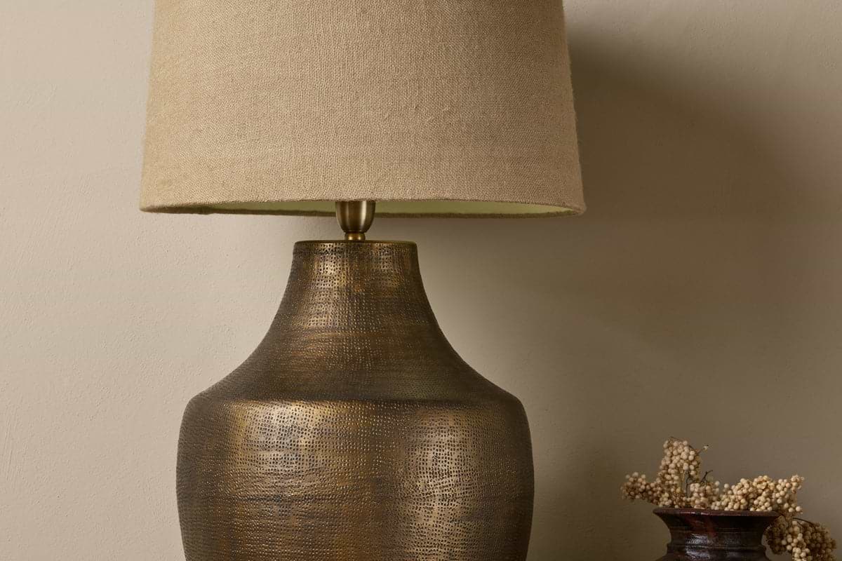 A close-up of the nkuku Tanda Etched Statement Table Lamp in Antique Brass (Extra Large) with a textured finish and beige fabric shade, set against a neutral wall. A small vase with dried flowers stands to the right.