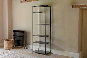 The Tanvi Display Shelf - Black - Narrow by nkuku stands against a beige wall. With five open shelves, it’s next to a gray radiator and woven basket, on tiled flooring with part of a rug beneath exposed wooden beams above.