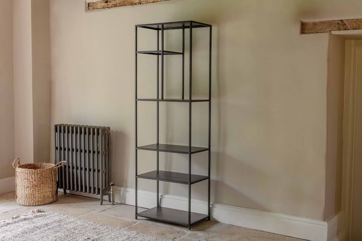The Tanvi Display Shelf - Black - Narrow by nkuku stands against a beige wall. With five open shelves, it’s next to a gray radiator and woven basket, on tiled flooring with part of a rug beneath exposed wooden beams above.