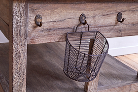 The nkuku Tivu Reclaimed Wood Butchers Block - Natural features a weathered finish, thick legs, lower shelf, and metal hooks beneath the tabletop. A small angled wire basket hangs from one hook, with side lighting highlighting the wood grain.