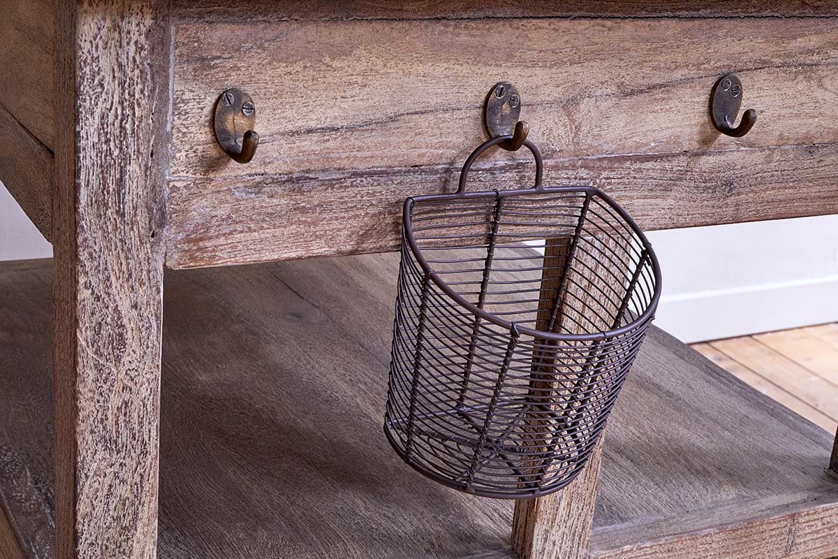 The nkuku Tivu Reclaimed Wood Butchers Block - Natural features a weathered finish, thick legs, lower shelf, and metal hooks beneath the tabletop. A small angled wire basket hangs from one hook, with side lighting highlighting the wood grain.