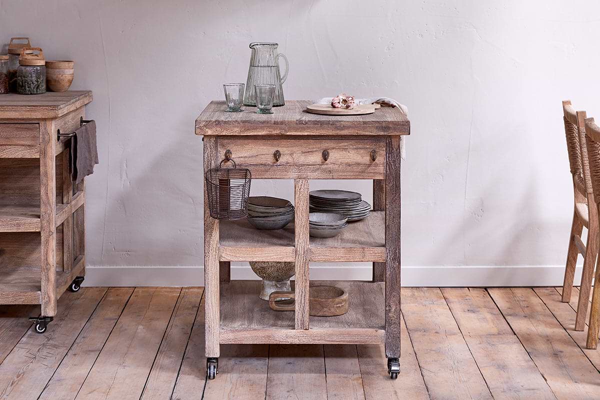 The nkuku Tivu Reclaimed Wood Butchers Block - Natural features shelves for dishes and bowls. A glass pitcher, cups, pastries, and a metal basket rest on top. The cart stands on a wooden floor with another cart and chairs nearby.