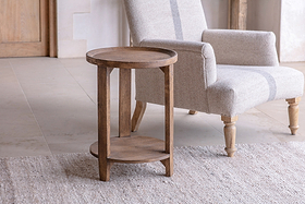 The nkuku Triveni Mango Wood Side Table in a walnut stain, with a lower shelf, stands on light tiled flooring beside a light gray upholstered armchair. A textured rug partially covers the floor in this softly lit, neutral room.