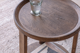 A glass of water sits on the nkuku Triveni Mango Wood Side Table - Walnut Stain, which features a round top with visible grain patterns, a raised edge, and sturdy wooden legs.