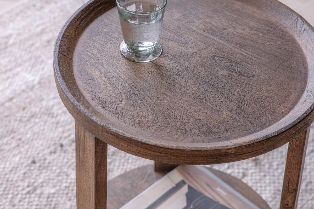 A glass of water sits on the nkuku Triveni Mango Wood Side Table - Walnut Stain, which features a round top with visible grain patterns, a raised edge, and sturdy wooden legs.