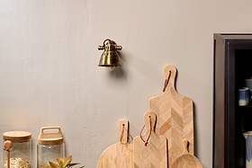 A Tubu Brass Spot Light by nkuku in antique brass casts a warm glow over glass jars with wooden lids, a small potted plant, and a black cabinet topped with stacked bowls. A brass wall bell with a brown leather strap hangs above light wooden cutting boards.