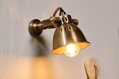 The nkuku Tubu Brass Spot Light in Antique Brass, inspired by industrial design, features a glowing bulb and vented shade. Its brown fabric cord casts warm yellow light and soft shadows on beige walls for a timeless touch.