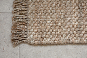 Close-up of nkuku’s Tunda Jute Doormat - Natural, featuring thick tan jute fibers and braided fringe, shown on a light gray stone tile floor. The textured doormat and floor tiles are both clearly visible.