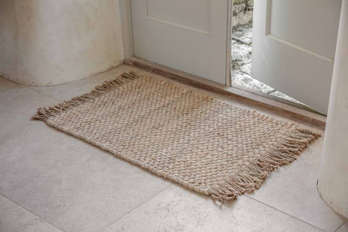 The nkuku Tunda Jute Doormat - Natural, featuring a chunky weave and fringed edges in beige jute, sits on a light stone tile floor by a slightly open white door letting in natural light.