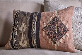 A rectangular woven pillow in earthy brown, beige, and tan with geometric diamonds and vertical stripes is styled with the nkuku Tussi Sia Cushion Cover in Dusty Pink, resting on a light sofa beside another textured neutral pillow.
