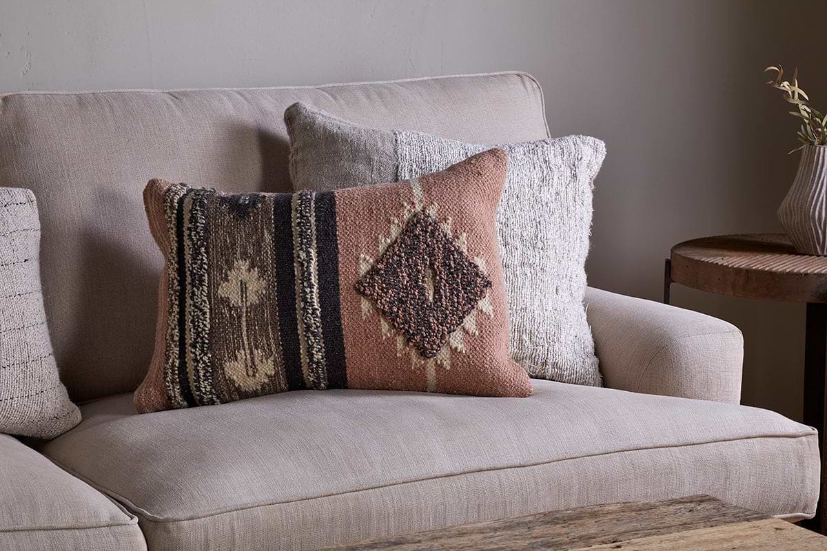 A beige sofa with two decorative pillows—one featuring the nkuku Tussi Sia Cushion Cover in dusty pink, the other patterned in earth tones. Feather inners add comfort, with a rustic wood table and small plant to the right.