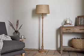 A cozy living room showcases the nkuku Usha Classic Wooden Floor Lamp - Natural, with a beige shade, positioned between a gray armchair with a draped blanket and a rustic wooden console table decorated with a basket and dried flowers.