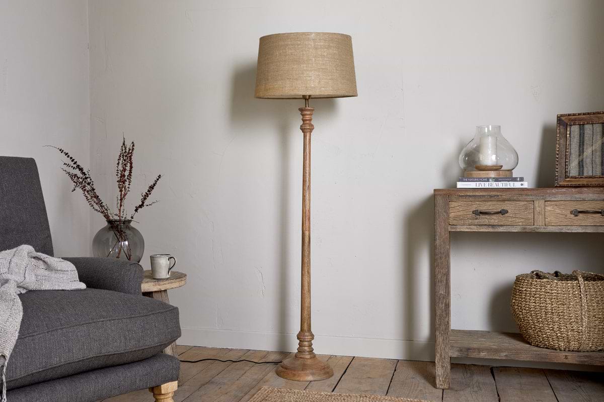 A cozy living room showcases the nkuku Usha Classic Wooden Floor Lamp - Natural, with a beige shade, positioned between a gray armchair with a draped blanket and a rustic wooden console table decorated with a basket and dried flowers.