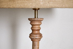 Close-up of nkuku's Usha Classic Wooden Floor Lamp - Natural, showcasing its hand-carved mango wood base with rounded details and metal rod, topped by a beige textured fabric shade against a light wall.