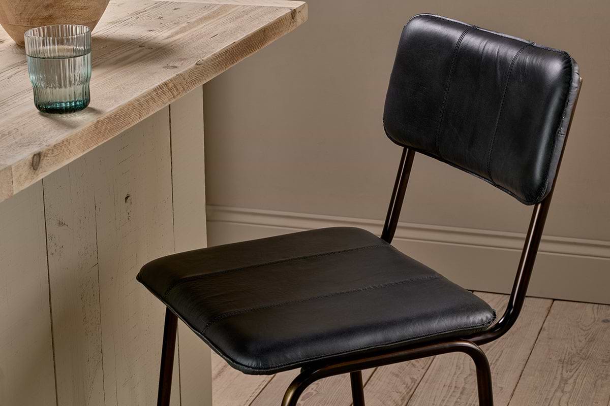 The nkuku Ukari Counter Chair in aged black leather with metal legs stands by a rustic wooden table. On the table are a clear ribbed glass and a round light brown item, set against light wood floors and neutral beige walls.
