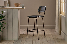 The nkuku Ukari Counter Chair - Aged Black, featuring a padded seat and backrest, stands on light wood flooring by a pale kitchen island with bowls and glasses as sunlight filters through sheer curtains near a green plant.