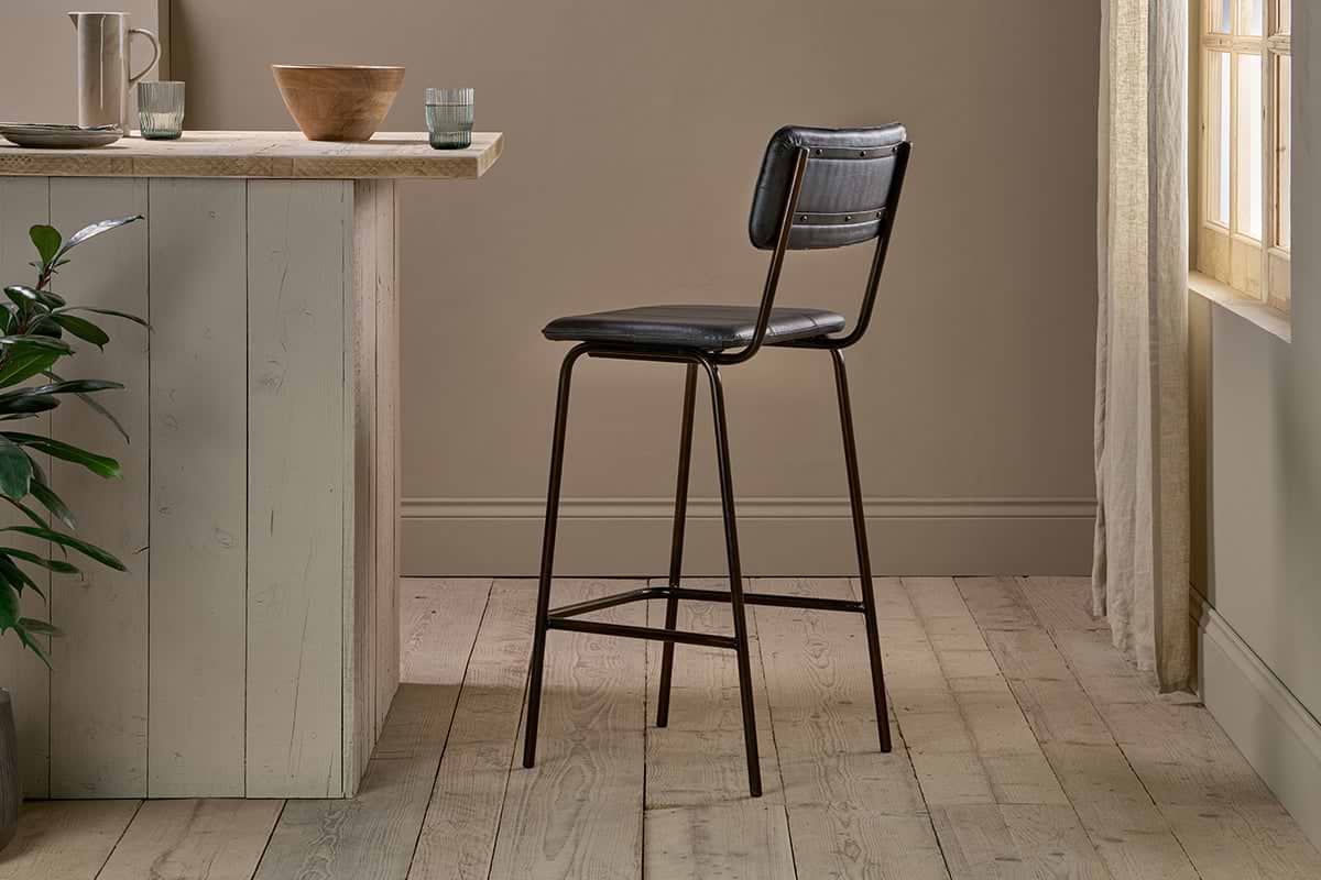 The nkuku Ukari Counter Chair - Aged Black, featuring a padded seat and backrest, stands on light wood flooring by a pale kitchen island with bowls and glasses as sunlight filters through sheer curtains near a green plant.