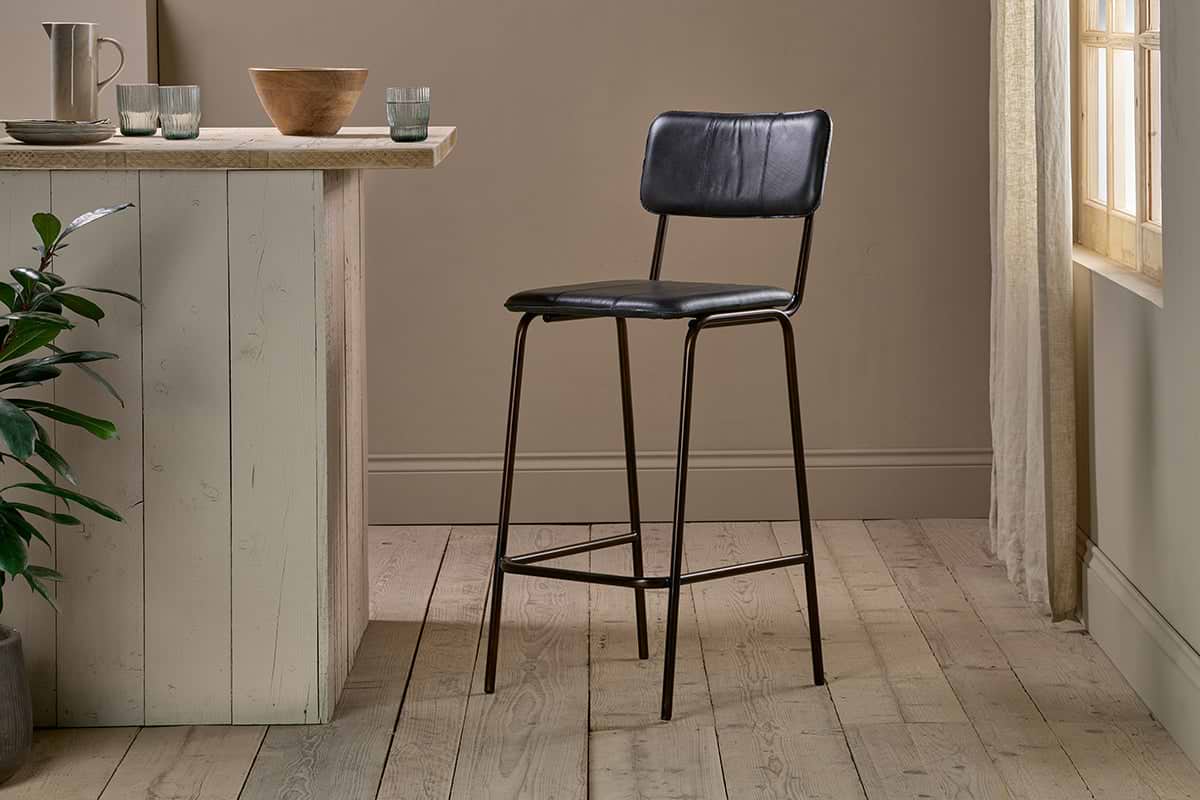 The nkuku Ukari Counter Chair in aged black leather and metal stands on light wooden flooring next to a rustic, light kitchen island. Sunlight through sheer curtains brightens the minimal, neutral space with simple decor.