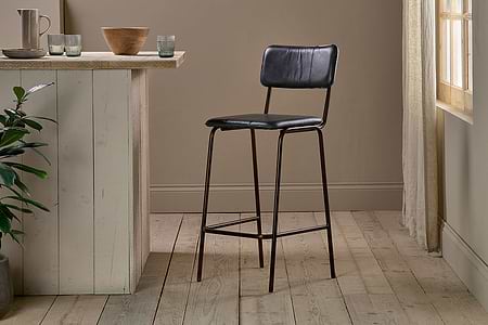 The nkuku Ukari Counter Chair in aged black leather and metal stands on light wooden flooring next to a rustic, light kitchen island. Sunlight through sheer curtains brightens the minimal, neutral space with simple decor.