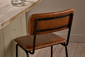 The nkuku Ukari Counter Chair - Aged Tan, featuring a handmade buffalo leather padded seat and backrest, stands by a rustic wooden table with a beige base. Two glasses of water sit on the table over light wood floors.