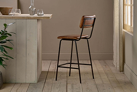 The nkuku Ukari Counter Chair - Aged Tan, with buffalo leather and a metal frame, is set beside a light wood kitchen island in a sunlit room with light wood floors, neutral walls, glassware, a plant, and sheer curtains.