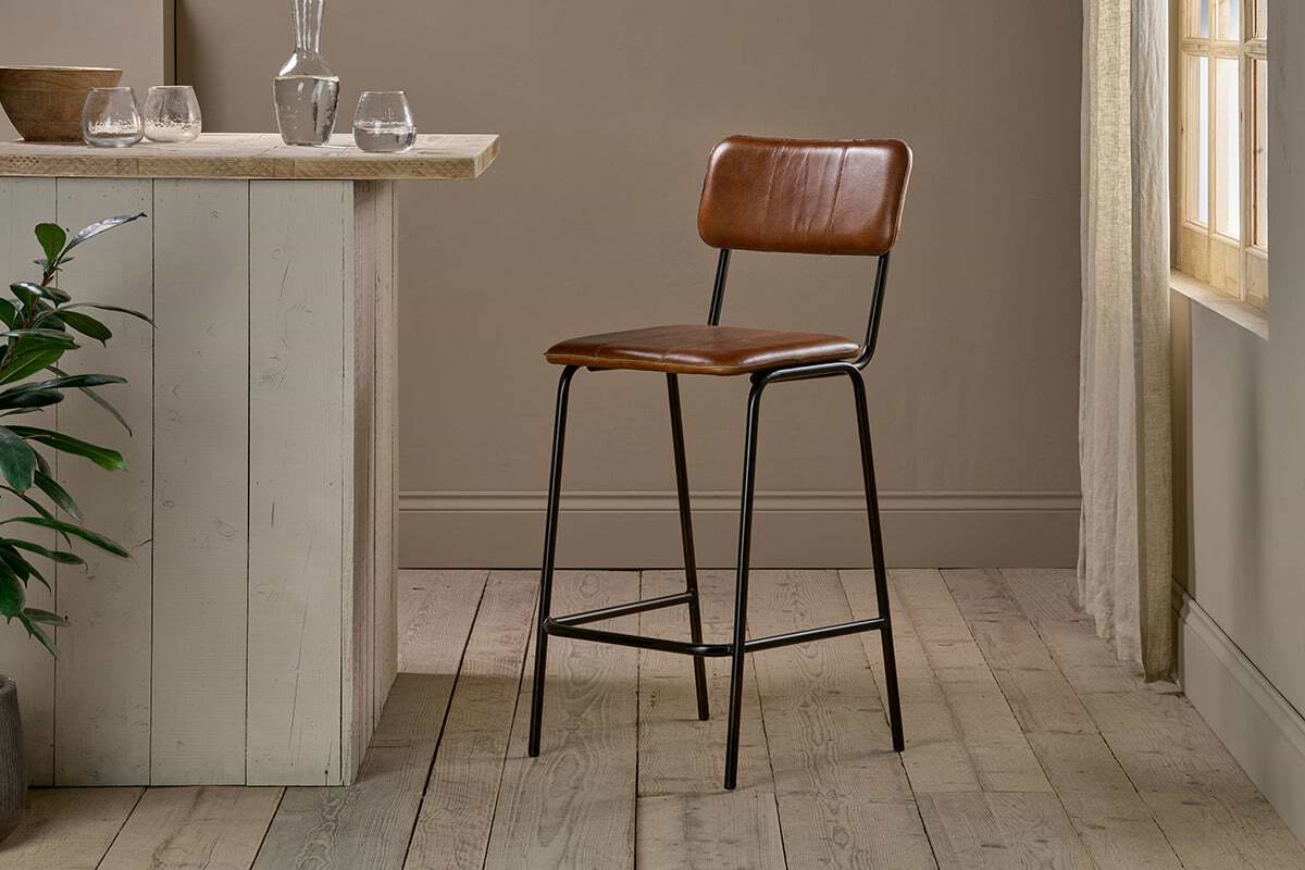 The nkuku Ukari Counter Chair - Aged Tan features a padded seat and back, slender black metal legs with footrest, standing by a rustic wooden counter in a neutral room with wood flooring and sunlight streaming in.