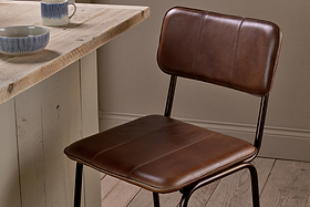 The nkuku Ukari Counter Chair in chocolate brown stands by a rustic wooden table. Two mugs and a patterned bowl sit atop the table, adding to the cozy, minimalist vibe of the wood-floored, neutral-walled space.