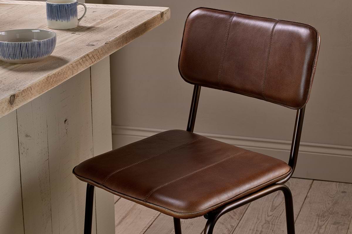 The nkuku Ukari Counter Chair in chocolate brown stands by a rustic wooden table. Two mugs and a patterned bowl sit atop the table, adding to the cozy, minimalist vibe of the wood-floored, neutral-walled space.