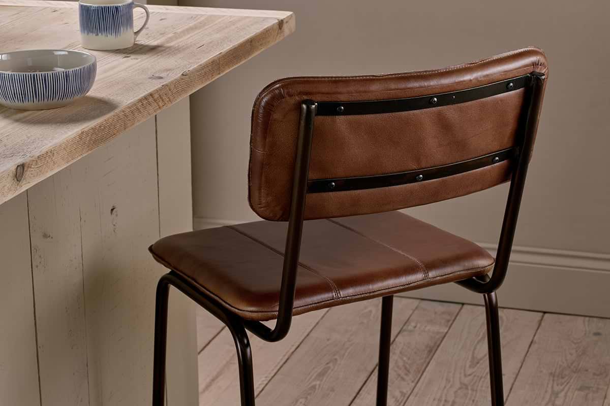 The Ukari Counter Chair - Chocolate Brown by nkuku, featuring black metal legs, is placed at a light wooden counter with blue and white patterned bowls and mugs, set against wood floors and neutral walls.