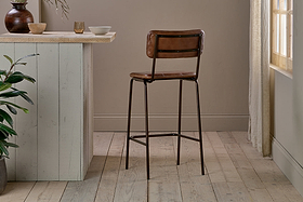 The nkuku Ukari Counter Chair in chocolate brown buffalo leather with slim black metal legs stands on light wood flooring by a pale wooden counter, accented by neutral bowls, a mug, and a plant as sunlight filters through a curtained window.