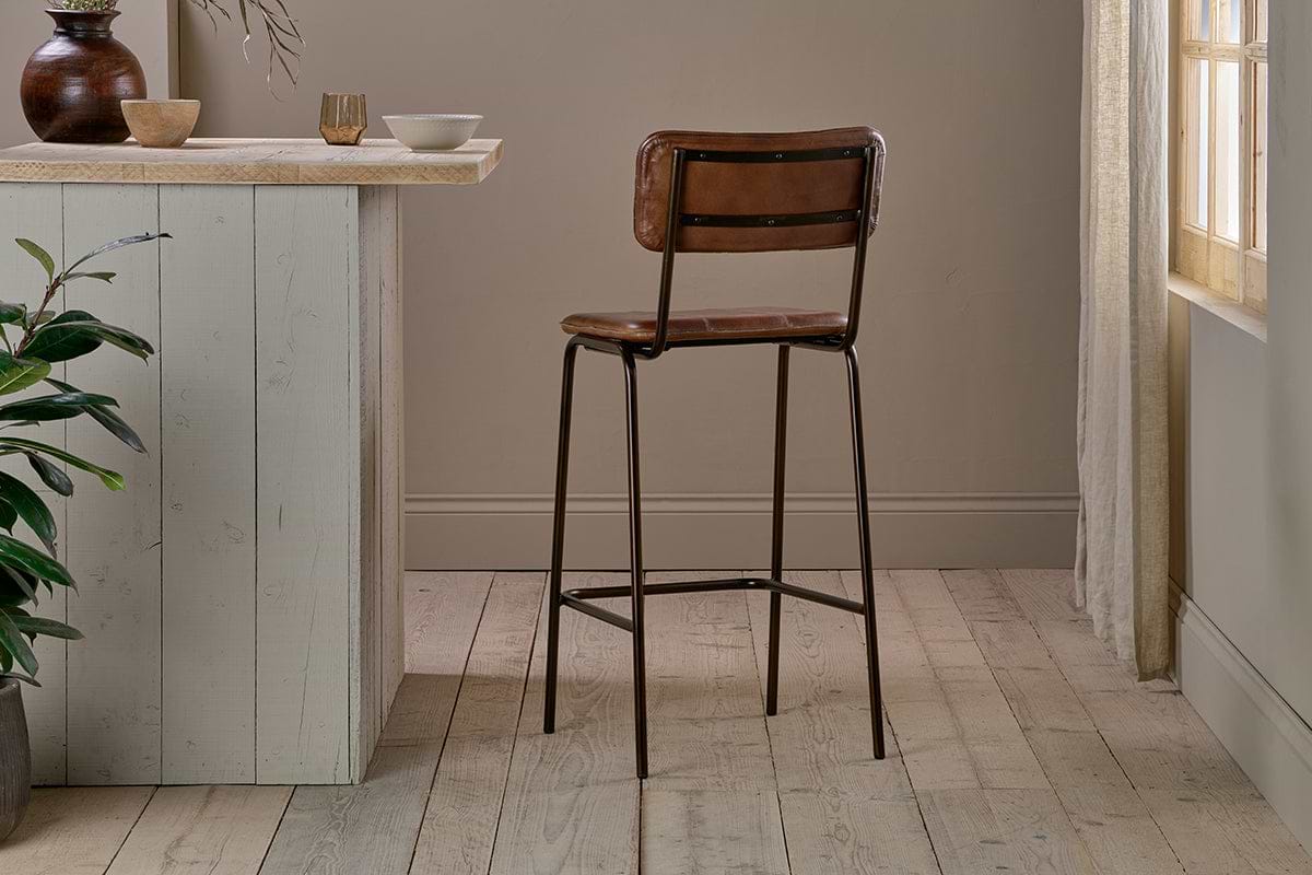 The nkuku Ukari Counter Chair in chocolate brown buffalo leather with slim black metal legs stands on light wood flooring by a pale wooden counter, accented by neutral bowls, a mug, and a plant as sunlight filters through a curtained window.
