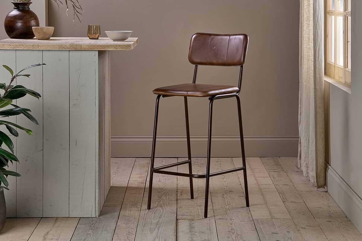 The nkuku Ukari Counter Chair - Chocolate Brown, featuring buffalo leather upholstery, stands on light wood flooring next to a pale gray counter with bowls, a cup, and a green plant beneath sunlit beige curtains.