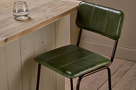 The nkuku Ukari Counter Chair in Rich Green, with a metal frame and leather upholstery, sits next to a rustic wood table. A round glass of water rests on the textured surface, surrounded by wooden floorboards and neutral walls.