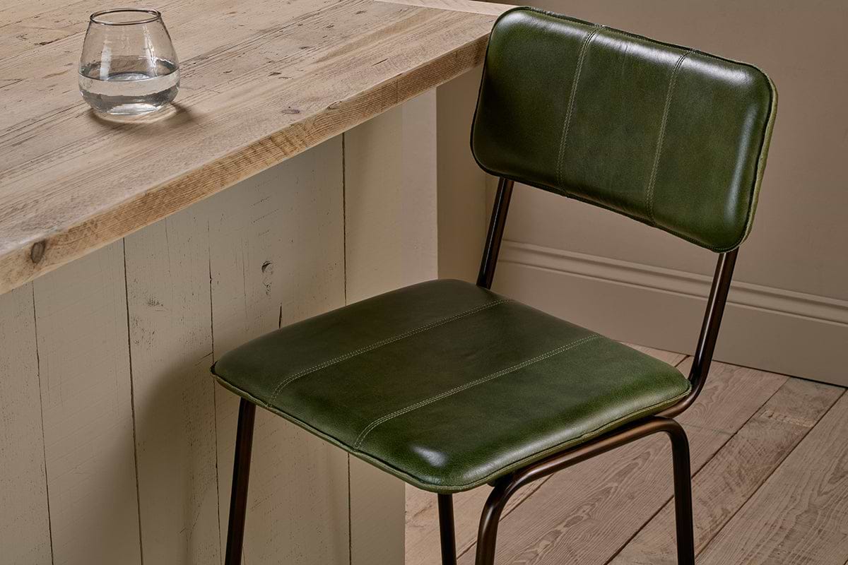 The nkuku Ukari Counter Chair in Rich Green, with a metal frame and leather upholstery, sits next to a rustic wood table. A round glass of water rests on the textured surface, surrounded by wooden floorboards and neutral walls.
