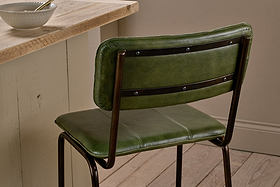 A nkuku Ukari Counter Chair in rich green leather, featuring a padded seat and backrest, sits by a light wood counter. On the wooden floor nearby, a white bowl with spoon complements the room’s neutral walls.